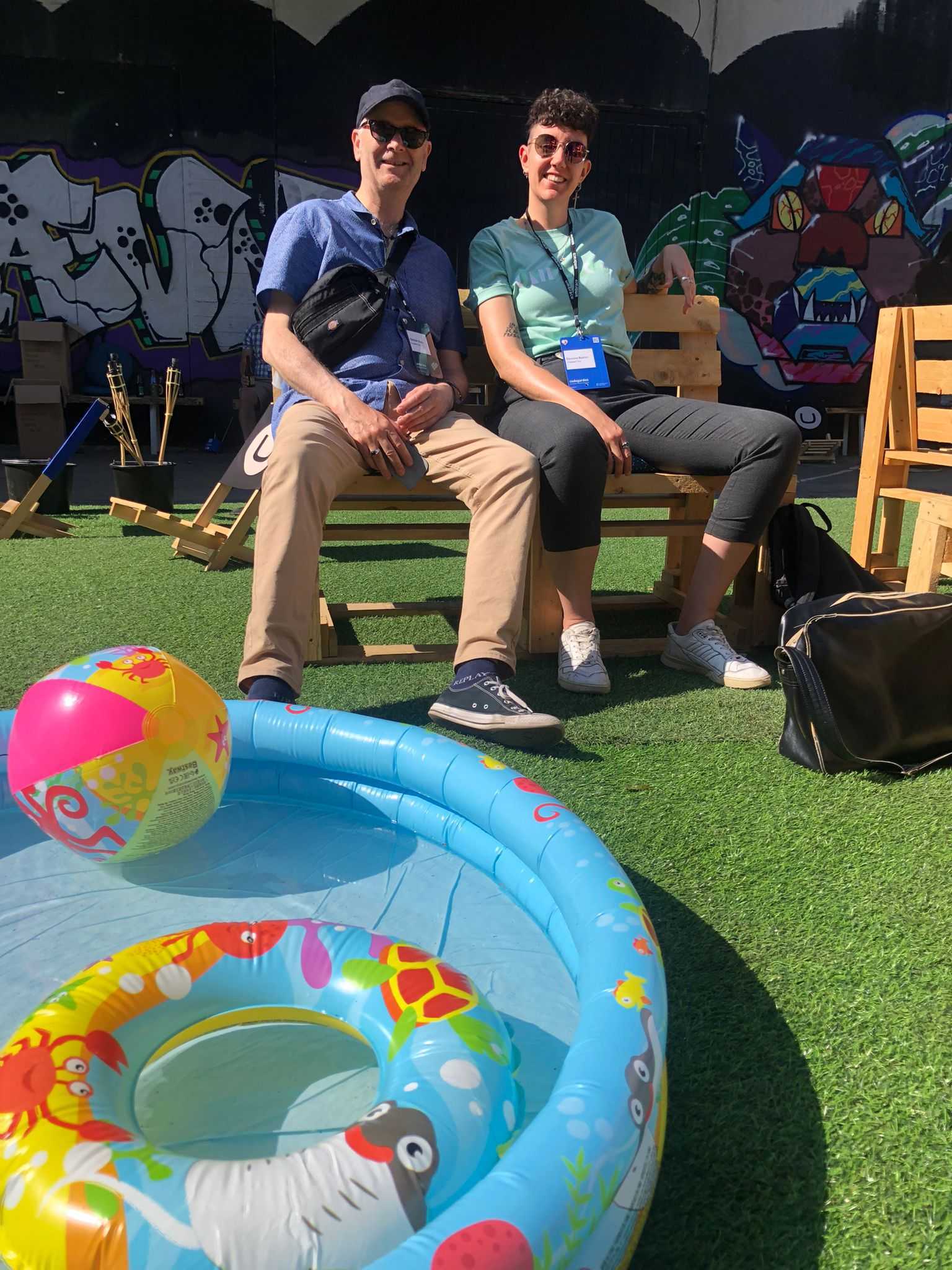 A picture of two people from Crumpled Dog sitting in the sun on a bench in the Codegarden outdoor section, next to a blue paddling pool with colourful inflatables. They are wearing sunglasses and smiling.