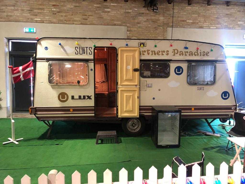Image of a stationary caravan inside a conference hall. There is a danish flag next to it, and it says "Partners Paradise" on the front.