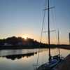 Image of a sailboat in the foreground on a canal, with the sun setting behind a row of houses and trees in the background