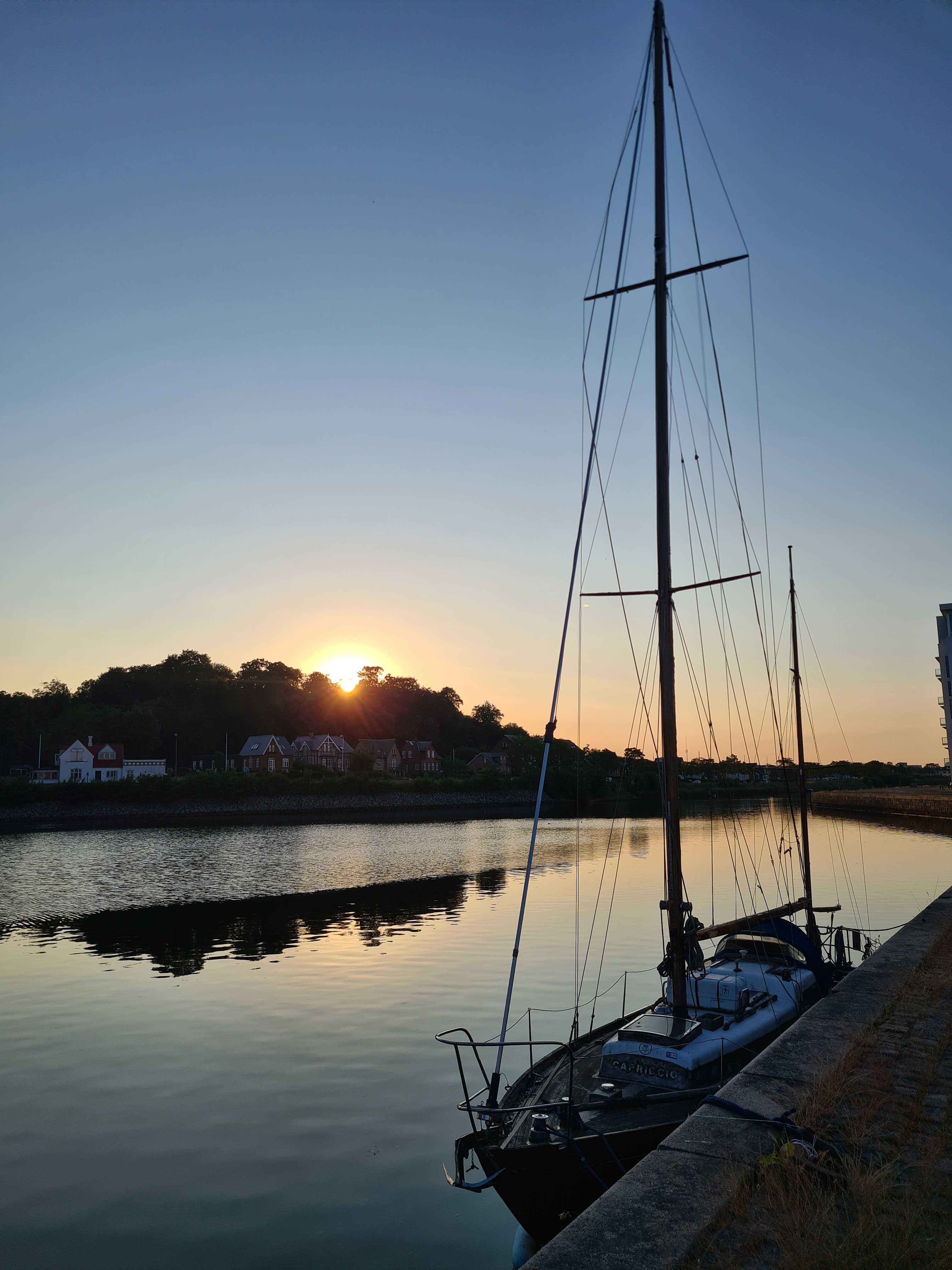 Image of a sailboat in the foreground on a canal, with the sun setting behind a row of houses and trees in the background