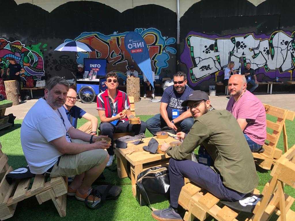 A group of six people from the Crumpled Dog team sitting in the sun around a picnic table, eating lunch. The walls in the background have graffiti on them, giving the impression of a festival.